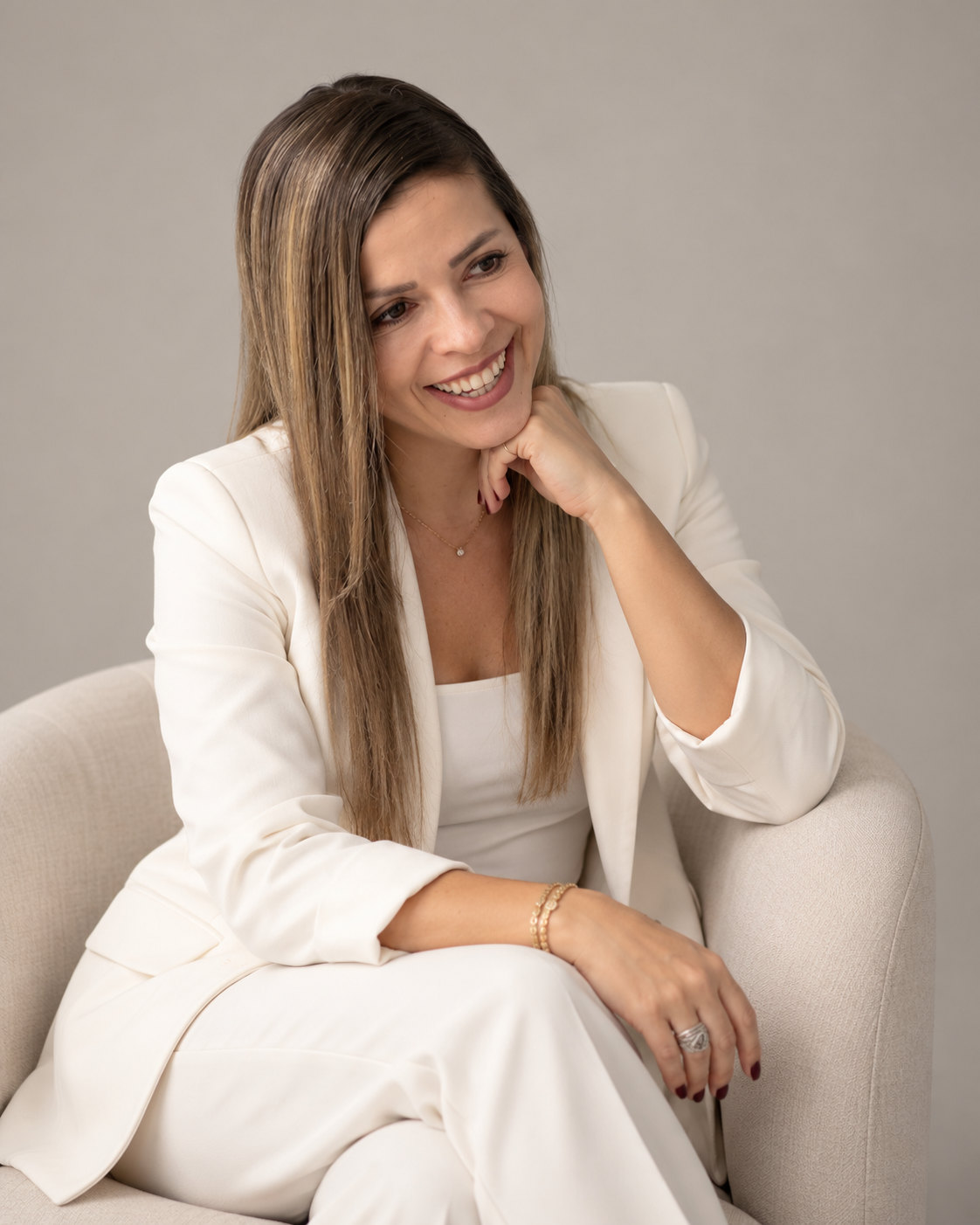 Alba Tejada V., sonriente, sentada en un sillón claro con traje blanco y la mano bajo el mentón