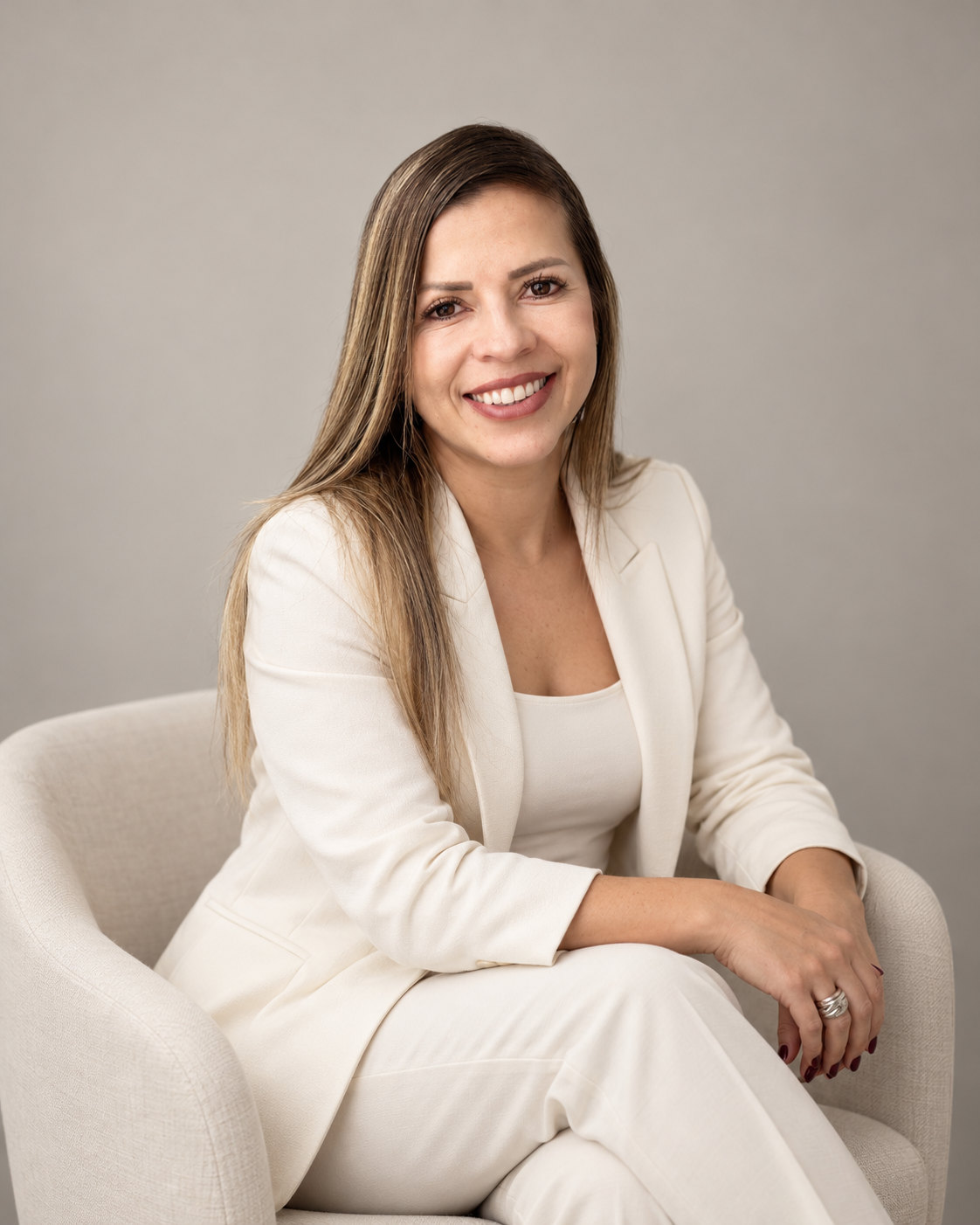 Retrato de Alba Tejada V. sonriendo a la cámara, sentada en un sillón claro con traje blanco