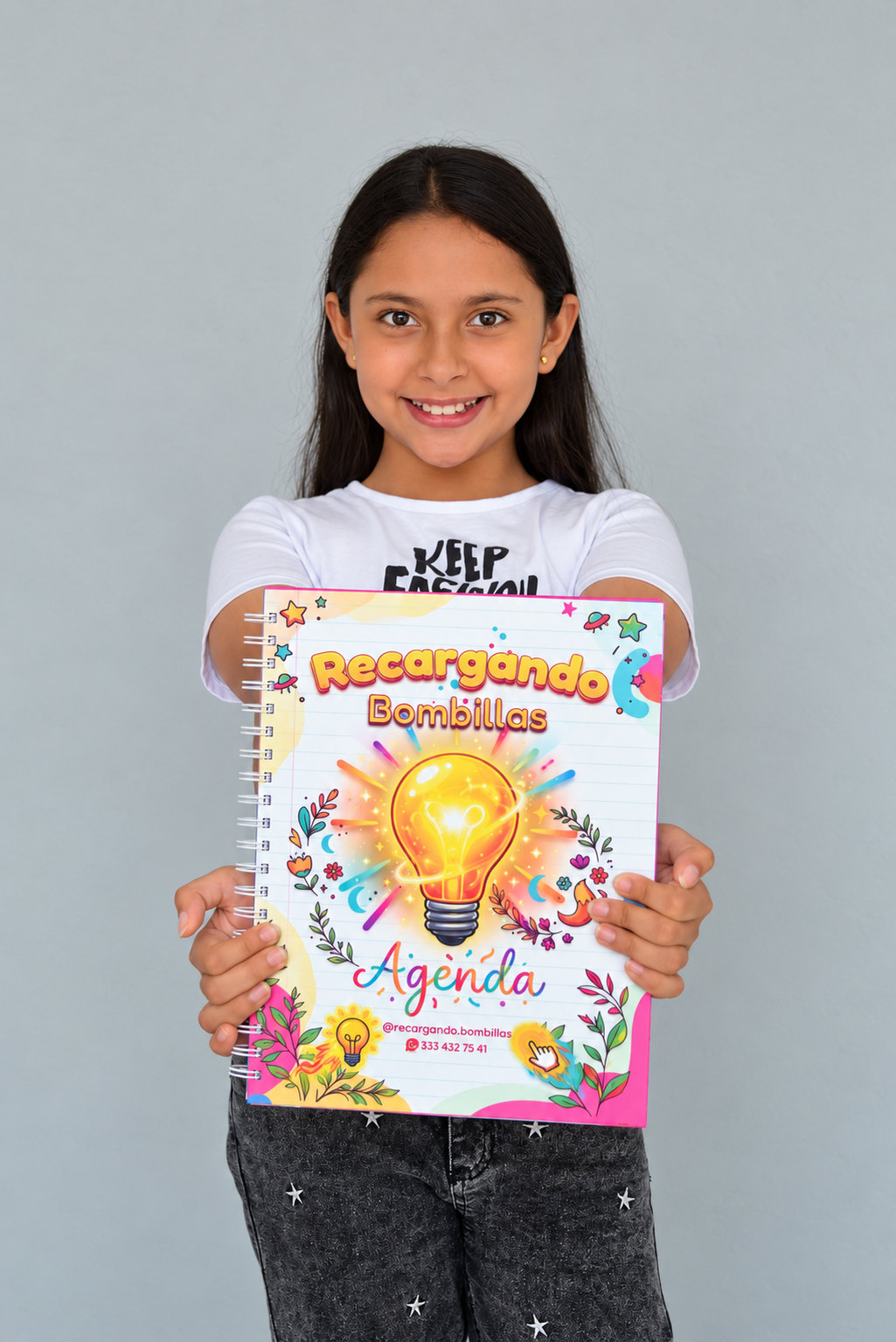 Niña sonriente sosteniendo hacia la cámara la agenda de Recargando Bombillas
