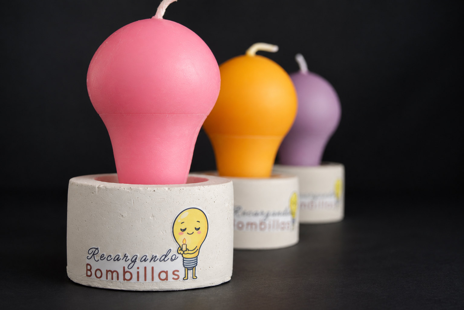Tres velas con forma de bombilla, rosada, naranja y lila, en bases de cemento con el logo de Recargando Bombillas